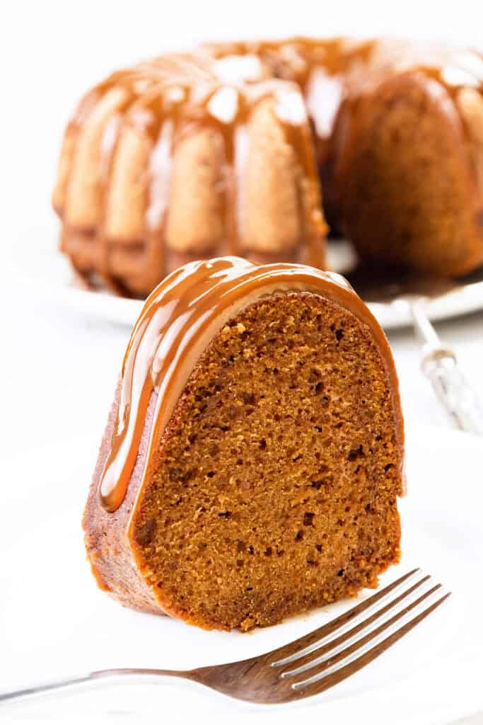 A slice of pumpkin bundt cake topped with caramel glaze sits on a white plate. The cake is moist and rich in texture, with a larger portion of the bundt cake visible in the background. A silver fork rests on the plate beside the slice.