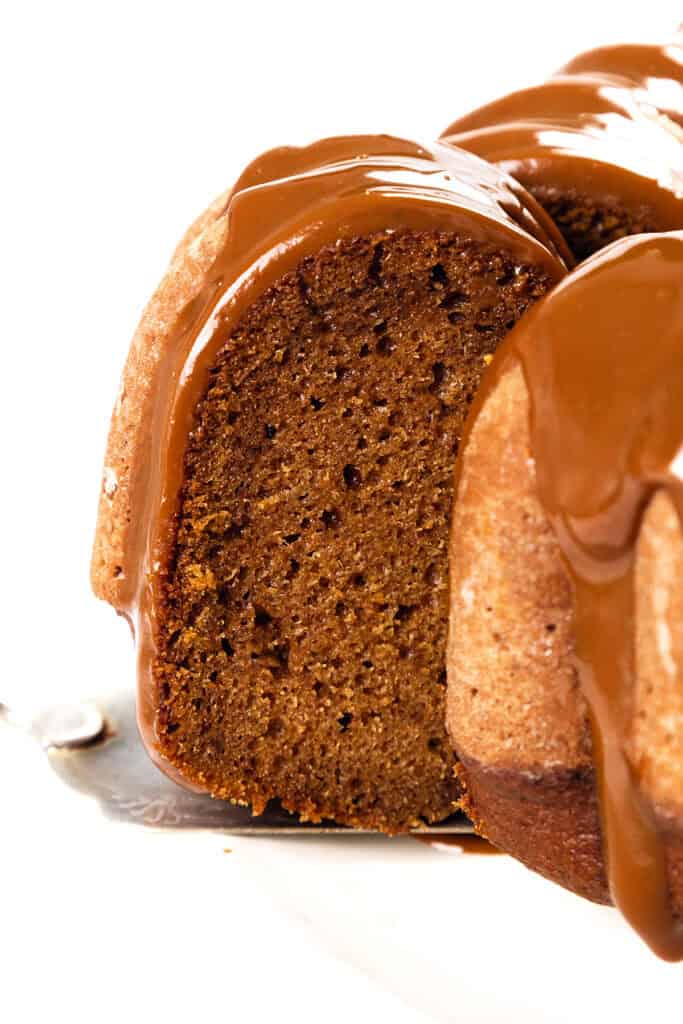 A close-up of a pumpkin bundt cake slice with rich brown coloring, drizzled with glossy caramel sauce. The cake's texture appears moist and dense, set on a metal cake server against a white background.