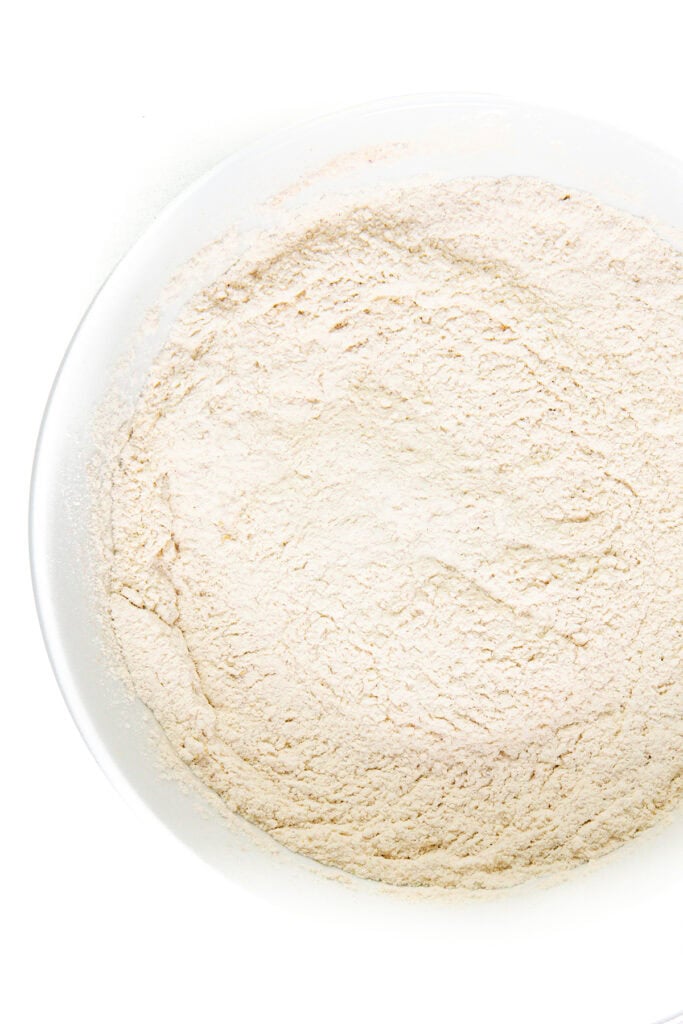 A close-up image of a white bowl filled with a fine, light beige powder, likely flour for a delightful Bundt cake, set against a white background. The texture appears smooth and evenly distributed across the bowl.