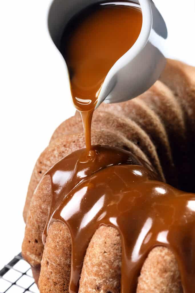 Close-up of a pumpkin bundt cake on a cooling rack, as rich caramel sauce gleams while cascading down its sides. The glaze highlights the cake's texture against a pristine white background.