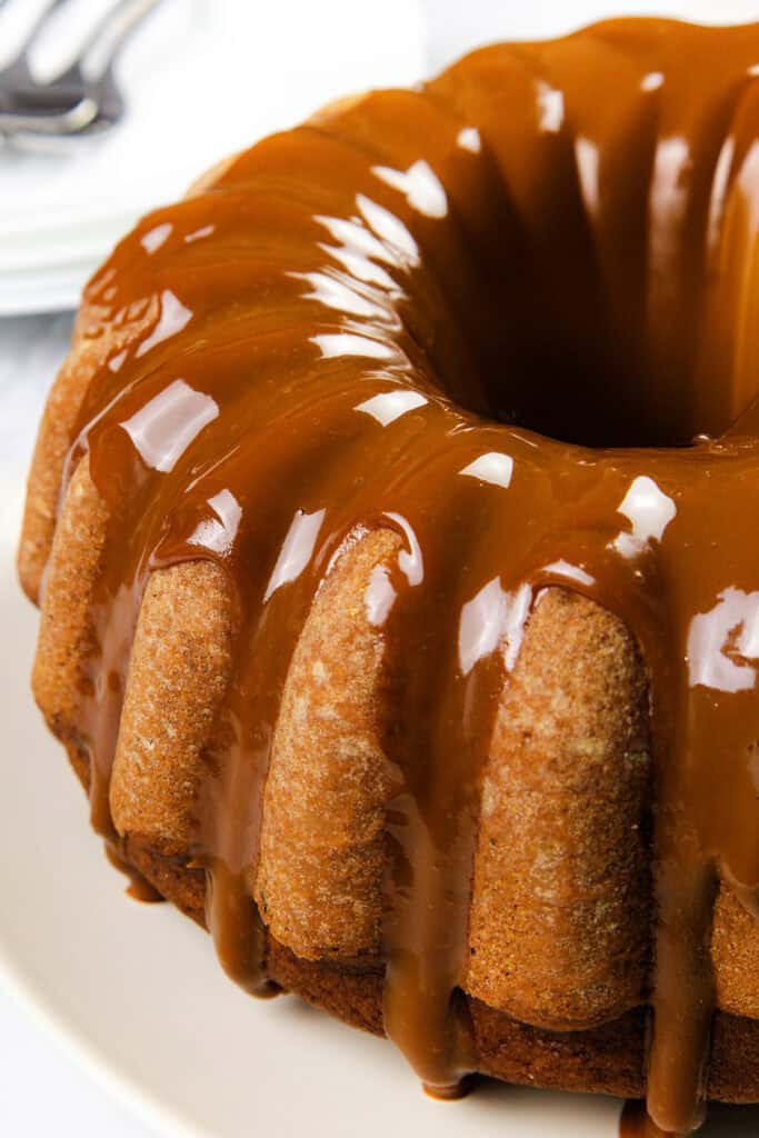 A close-up of a pumpkin bundt cake drizzled with glossy caramel sauce, set on a white plate. The caramel cascades down the cake's ridges, creating a rich, inviting appearance. In the background, there's a hint of stacked white plates.