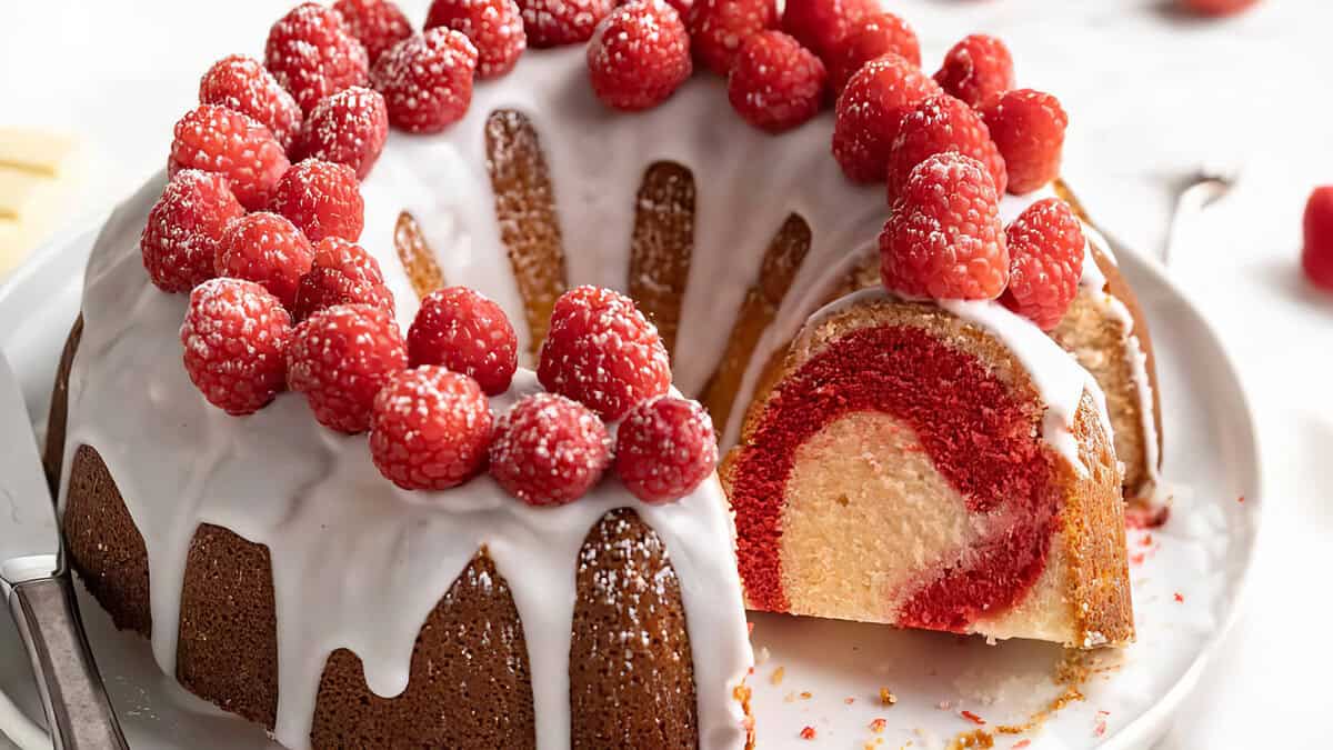 A sliced bundt cake, reminiscent of popular bundt cake recipes, features a red swirl pattern inside and is adorned with white icing and fresh raspberries. Some berries are lightly dusted with sugar. A knife rests beside the cake on a pristine white plate.