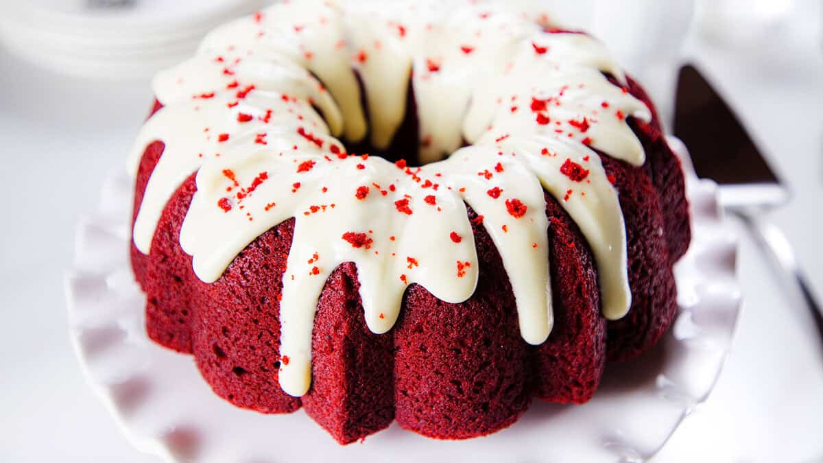 A red velvet bundt cake topped with a thick layer of white cream cheese frosting and sprinkled with red cake crumbs, displayed on a white scalloped cake stand.