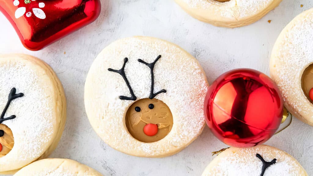 Festive cookies decorated with reindeer faces, featuring antlers and red noses, make for delightful holiday treats. Each cookie is dusted with powdered sugar, perfected from cherished cookie recipes. A shiny red Christmas ornament is placed nearby on a white surface.