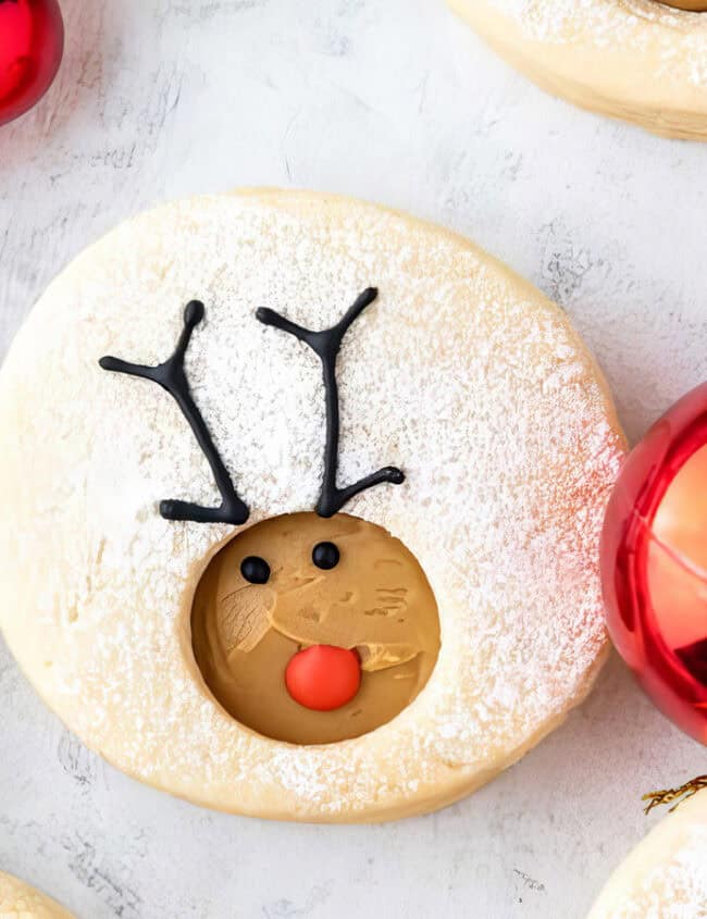 Festive cookies decorated with reindeer faces, featuring antlers and red noses, make for delightful holiday treats. Each cookie is dusted with powdered sugar, perfected from cherished cookie recipes. A shiny red Christmas ornament is placed nearby on a white surface.