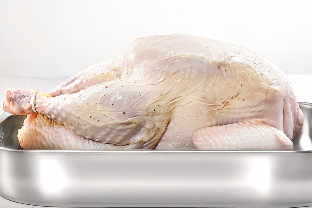 A raw, whole turkey sits in a stainless steel roasting pan against a white background. The pale, textured skin is gently seasoned, hinting at the delicious transformation into a slow-roasted turkey.