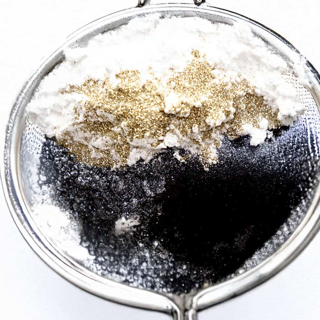 A shiny sieve holds a blend of white flour, black cocoa powder, and gold glitter, creating a striking contrast. 