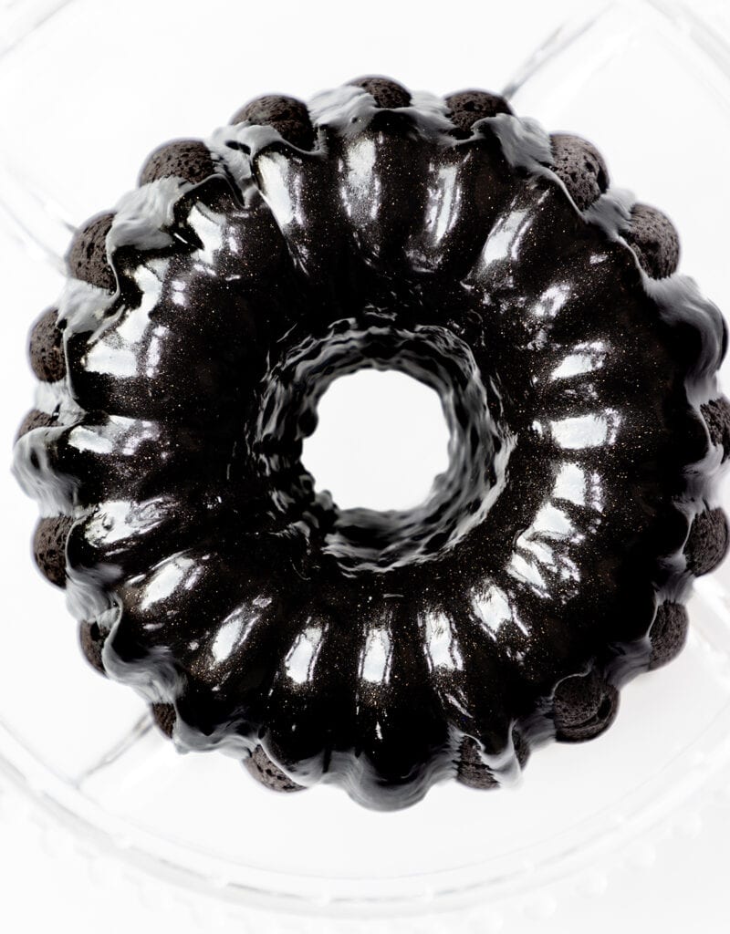 A glossy black bundt cake with a shiny glaze is displayed on a clear glass stand. The cake's intricate ridges are visible, creating an elegant and appealing presentation against the white background.