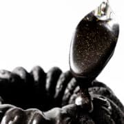 A spoon drizzles glossy, black glaze with golden specks over a ridged black cake adorned with a silky black cream cheese glaze against a white background.