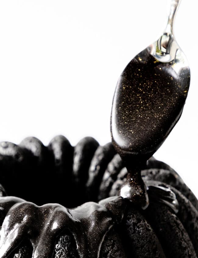 A spoon drizzles glossy, black glaze with golden specks over a ridged black cake adorned with a silky black cream cheese glaze against a white background.