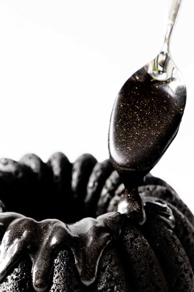 A spoon drizzles glossy, black glaze with golden specks over a ridged black cake adorned with a silky black cream cheese glaze against a white background.