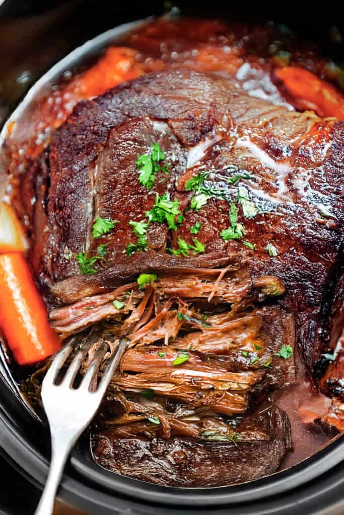 A tender, slow-cooked Crock Pot sirloin tip roast with shredded beef in a rich, savory sauce. Garnished with chopped parsley, the roast is accompanied by chunks of carrots. A fork is placed in the meat, highlighting its juicy, fall-apart texture.