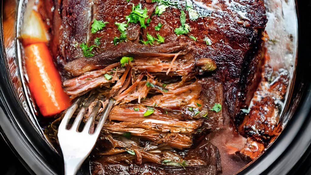 A close-up of a slow-cooked sirloin tip roast in a crockpot. The tender meat is shredded and garnished with fresh parsley, accompanied by a large carrot. A fork is placed within the roast, showing its tenderness and juiciness.