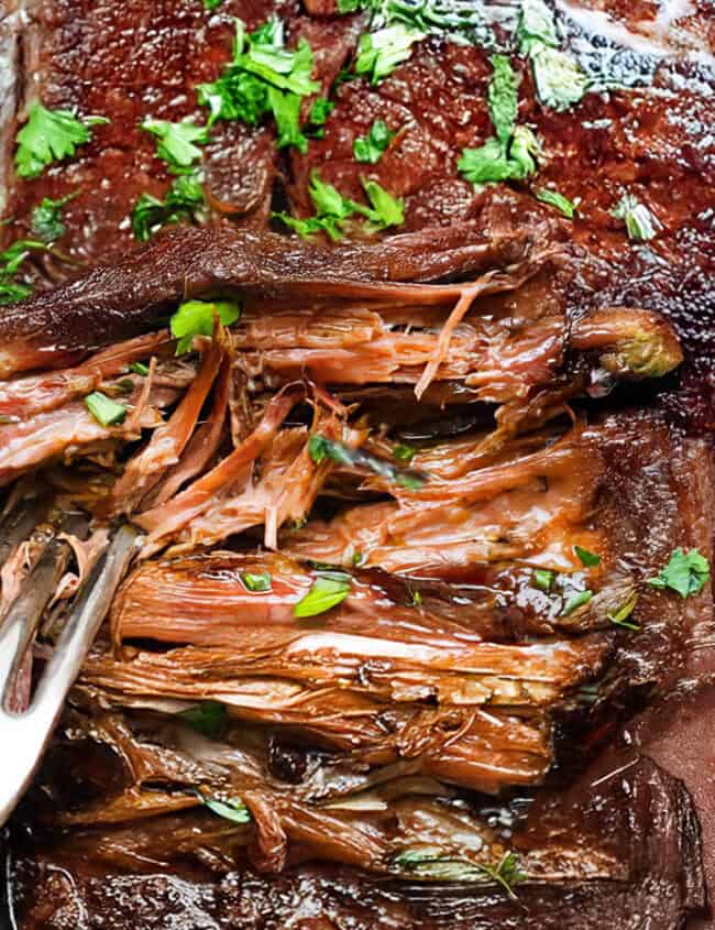 A close-up of a slow-cooked sirloin tip roast in a crockpot. The tender meat is shredded and garnished with fresh parsley, accompanied by a large carrot. A fork is placed within the roast, showing its tenderness and juiciness.