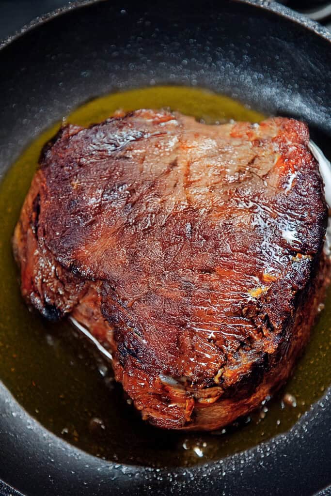 A seared Sirloin Tip Roast sits in a black skillet, glistening with oil. The meat is browned with a slightly crispy crust, indicating it was cooked to create a flavorful outer layer. The skillet shows a small amount of oil pooled around the roast.