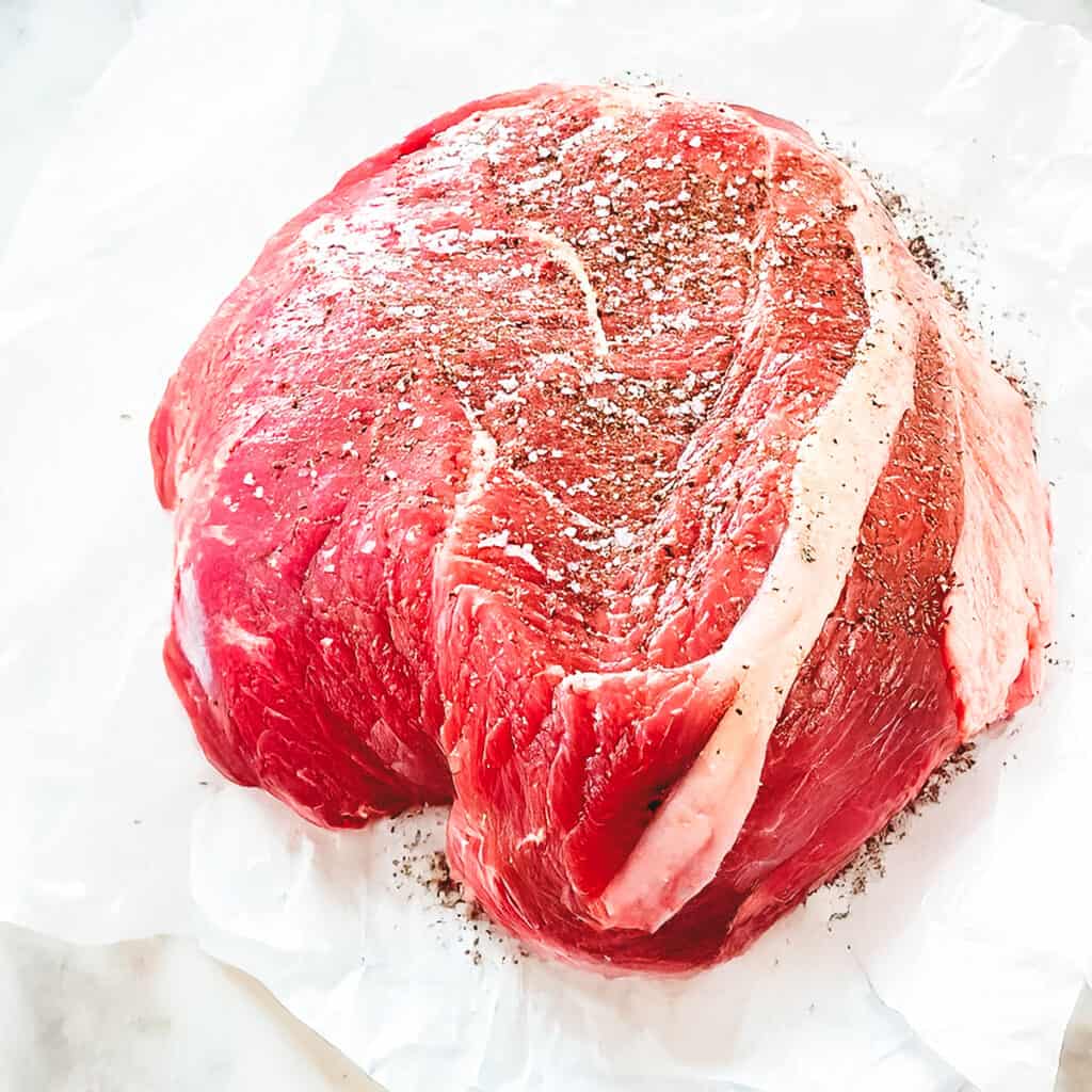 An uncooked sirloin tip roast with marbling of fat sits on crinkled white parchment paper. The beef is lightly sprinkled with salt and pepper, highlighting the texture and color of the meat. Ideal for a crock pot recipe, the background is a soft, neutral-toned surface.