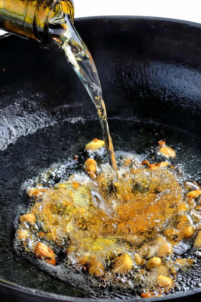 Oil being poured from a bottle into a cast iron skillet with sautéing garlic cloves. The golden brown garlic sizzles as the hot oil bubbles around it. Meanwhile, in the background, a Crock Pot roast simmers slowly, infusing the kitchen with rich aromas.