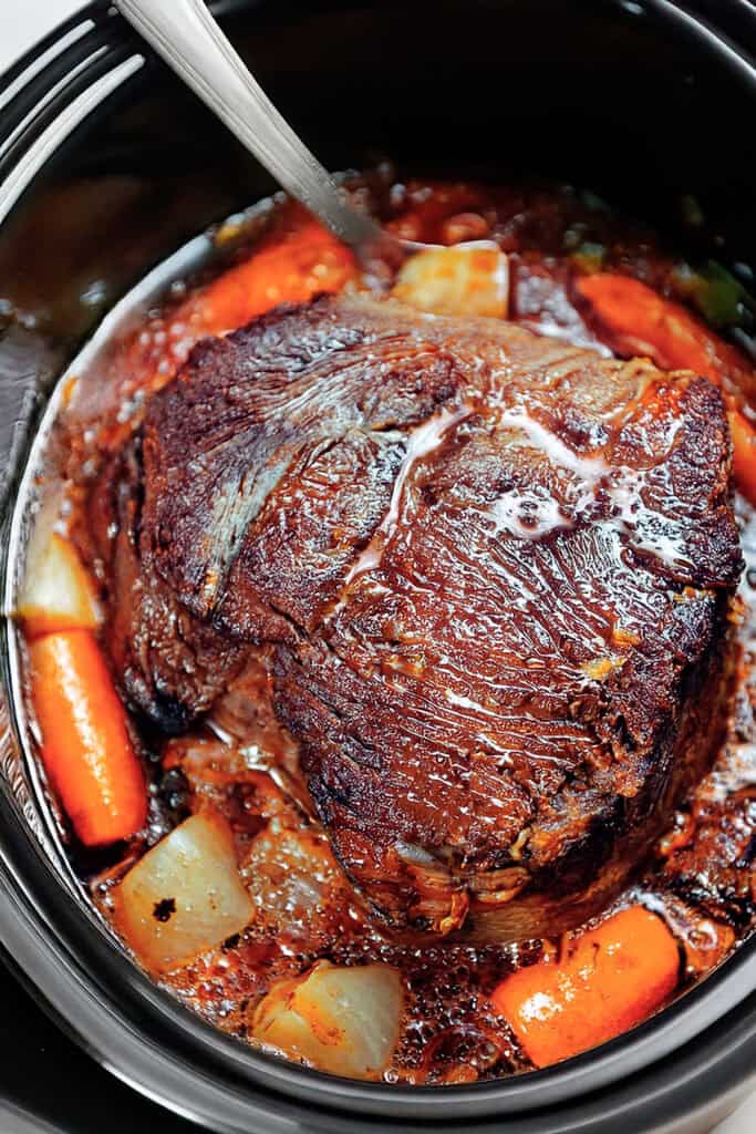 A juicy sirloin tip roast sits in a slow cooker, surrounded by tender carrots and onions in a rich, brown broth. A serving spoon rests on the edge.