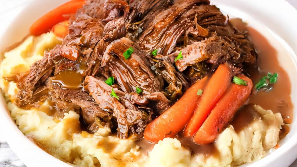 A plate containing shredded sirloin tip pot roast served on top of mashed potatoes, garnished with baby carrots and fresh parsley. The dish is accompanied by a savory brown gravy.