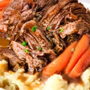 A plate containing shredded sirloin tip pot roast served on top of mashed potatoes, garnished with baby carrots and fresh parsley. The dish is accompanied by a savory brown gravy.