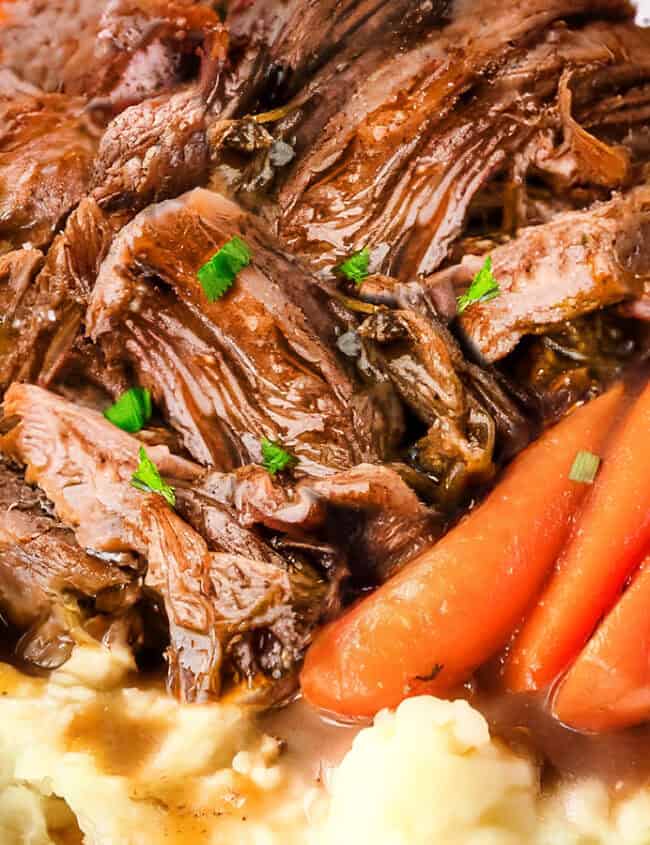 A plate containing shredded sirloin tip pot roast served on top of mashed potatoes, garnished with baby carrots and fresh parsley. The dish is accompanied by a savory brown gravy.