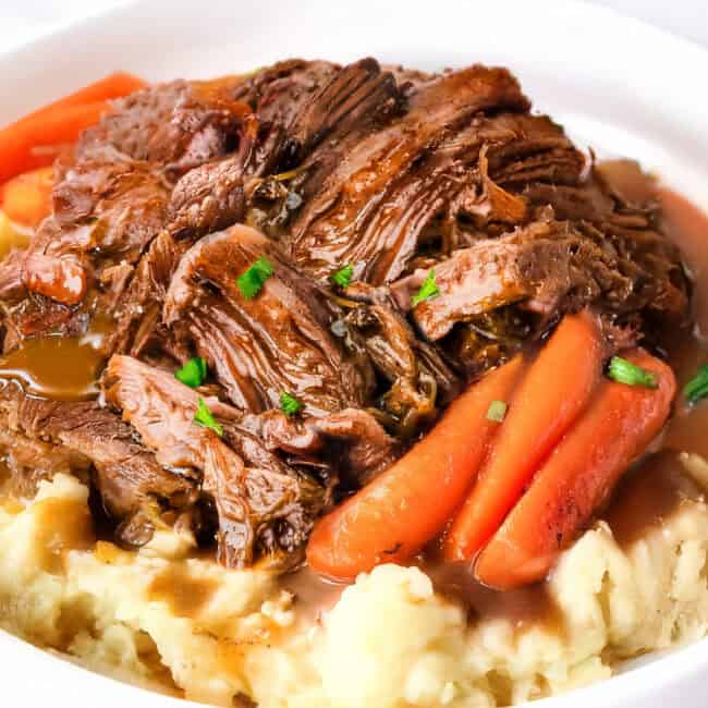 A white bowl cradles a Crock Pot sirloin tip roast, tenderly shredded over creamy mashed potatoes. Carrot pieces are nestled around the beef, garnished with chopped green herbs, all resting on a blue napkin.