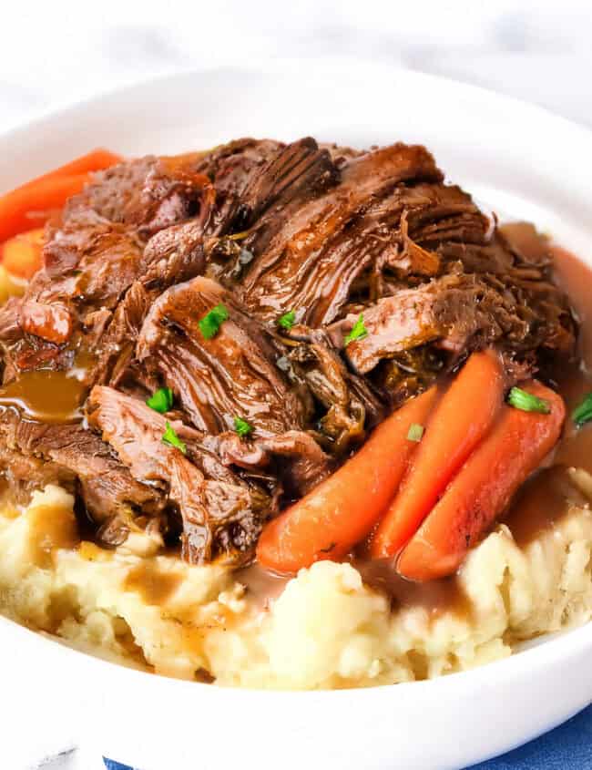 A white bowl cradles a Crock Pot sirloin tip roast, tenderly shredded over creamy mashed potatoes. Carrot pieces are nestled around the beef, garnished with chopped green herbs, all resting on a blue napkin.