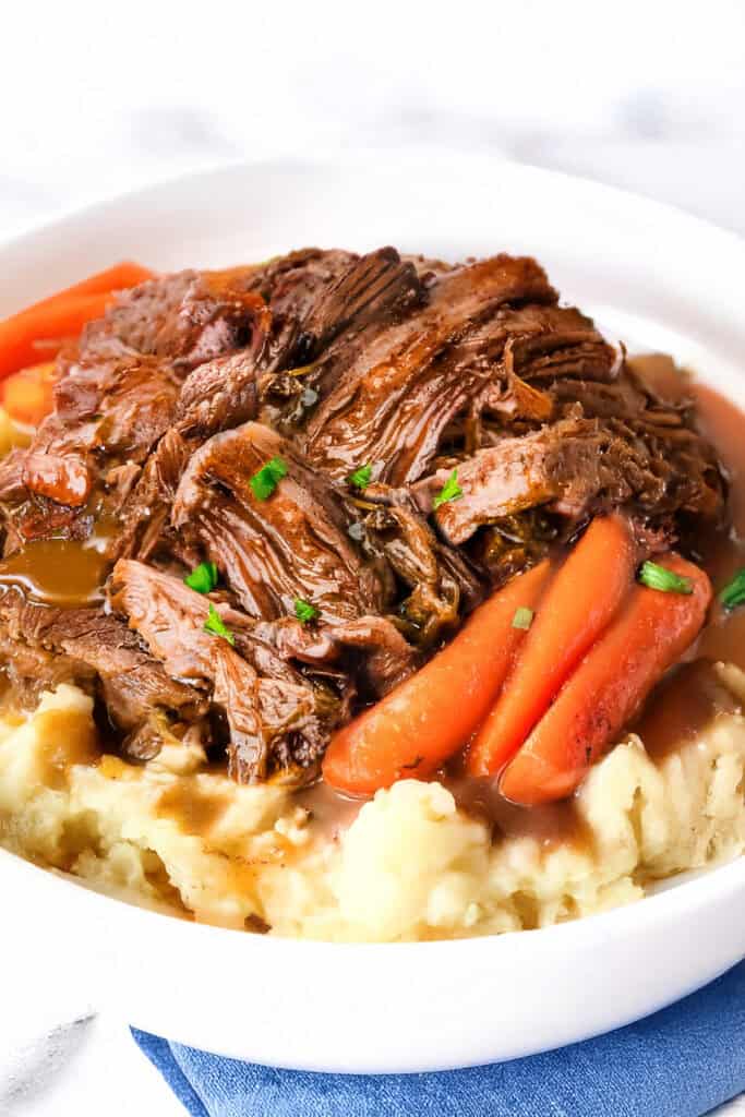 A white bowl cradles a Crock Pot sirloin tip roast, tenderly shredded over creamy mashed potatoes. Carrot pieces are nestled around the beef, garnished with chopped green herbs, all resting on a blue napkin.