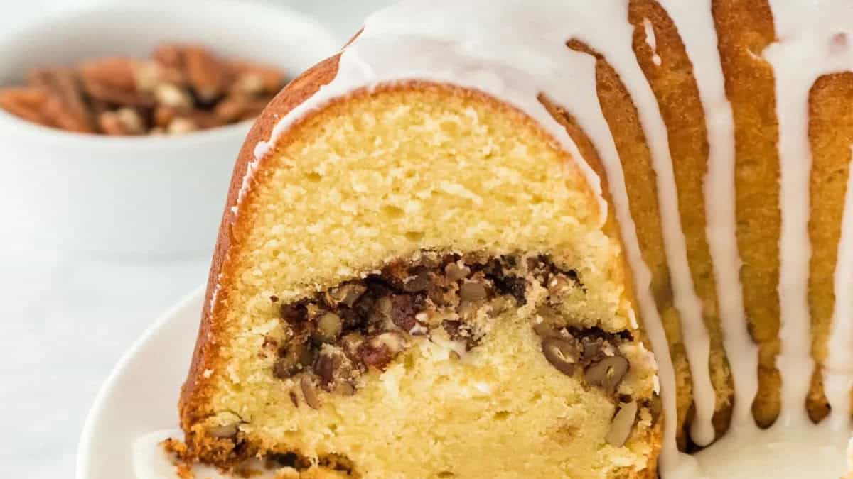 A sliced bundt cake topped with white icing reveals its rich, moist interior filled with chopped pecans. Perfect for those exploring Bundt Cake Recipes, a bowl of pecans blurs softly in the background on a pristine white surface.