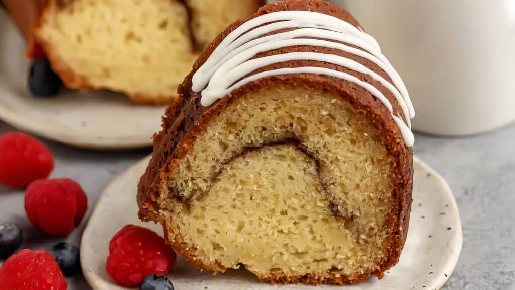 Close-up of a slice of bundt cake with a swirl of icing on top. The cake is on a ceramic plate, surrounded by fresh raspberries and blueberries. A larger piece of cake is partially visible in the background on another plate.