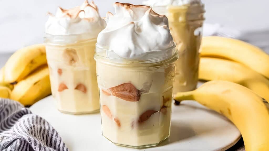 Glass jars filled with Southern-style banana pudding, garnished with whipped cream, surrounded by whole bananas on a white plate. A striped cloth is placed nearby.