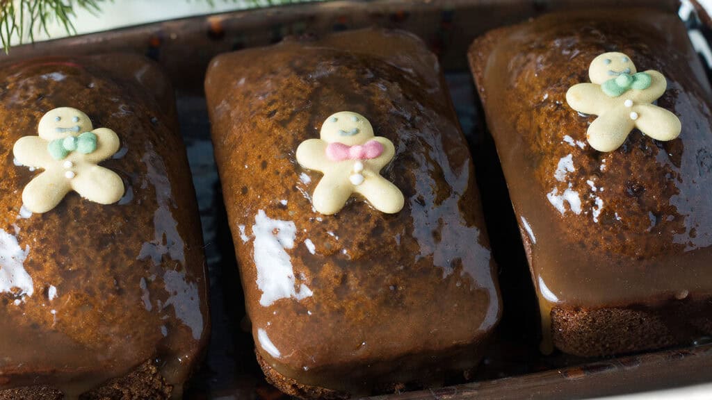 Three glazed gingerbread loaves, topped with tiny gingerbread men decorations, each adorned with colorful candy ties. These irresistible desserts are arranged in a row on a tray—perfect for those who simply can't get enough of holiday treats.