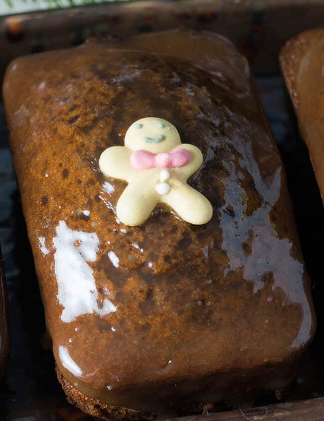 Three glazed gingerbread loaves, topped with tiny gingerbread men decorations, each adorned with colorful candy ties. These irresistible desserts are arranged in a row on a tray—perfect for those who simply can't get enough of holiday treats.