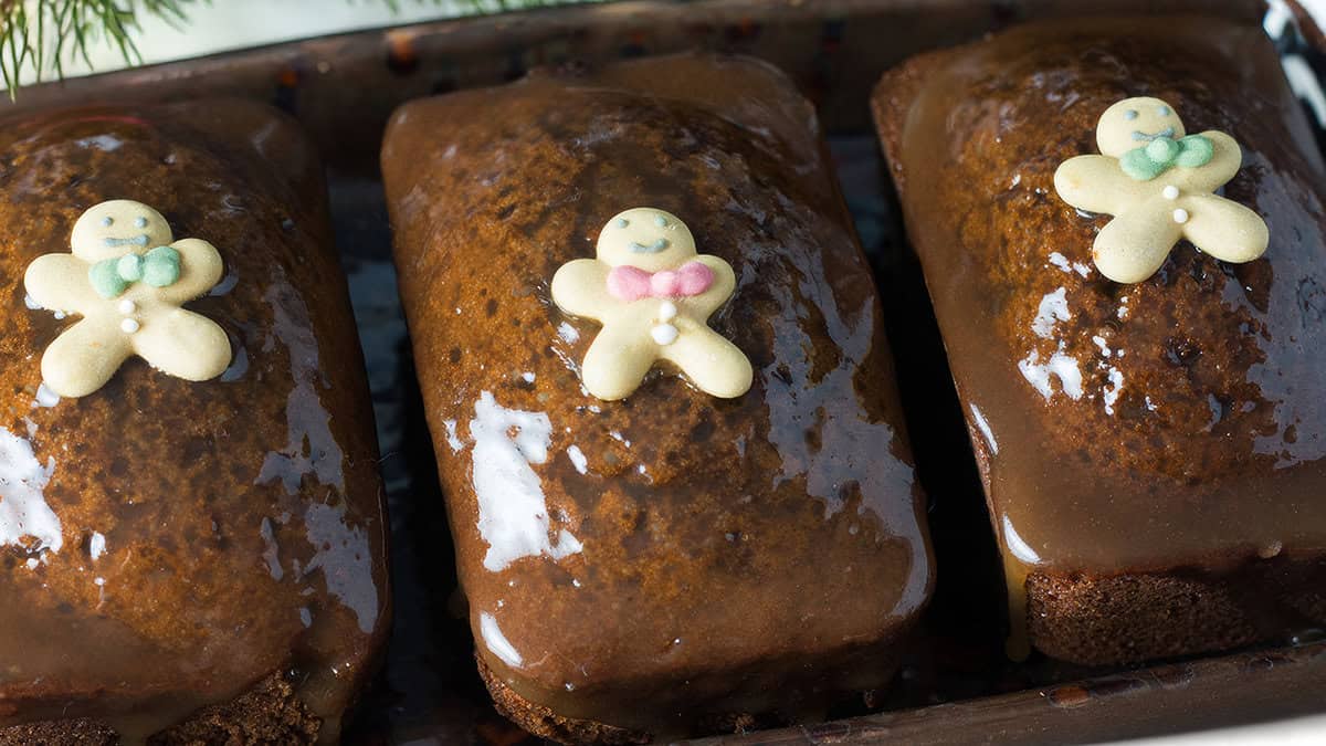 Three rectangular gingerbread cakes, topped with a shining caramel glaze and decorated with tiny gingerbread men sporting colored icing accents, are arranged side by side on a tray. These desserts are irresistibly delightful—you simply can't get enough.