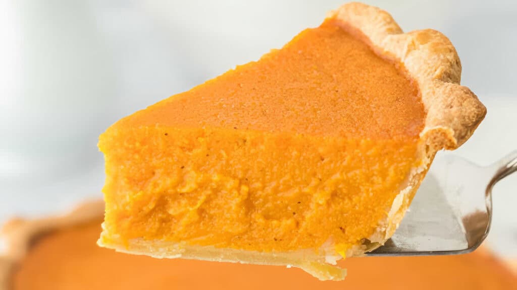 A close-up of a slice of pumpkin pie being held up with a silver spatula. The pie has a smooth, orange filling and a golden-brown crust, set against a soft-focus background.