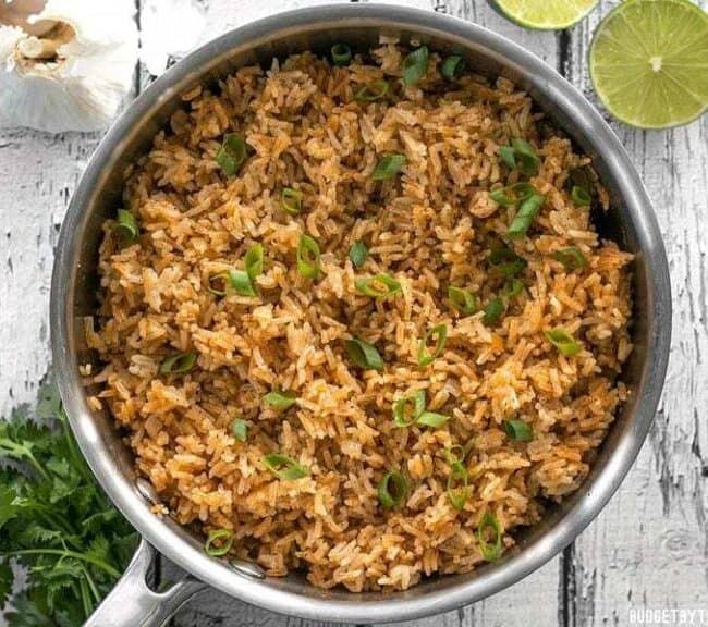 A silver pot filled with seasoned rice, perfect for rice dish recipes, is garnished with chopped green onions. Placed on a weathered wooden surface, cilantro, garlic, and sliced limes are nearby. The fluffy rice looks flavorful, integrated with herbs and spices.