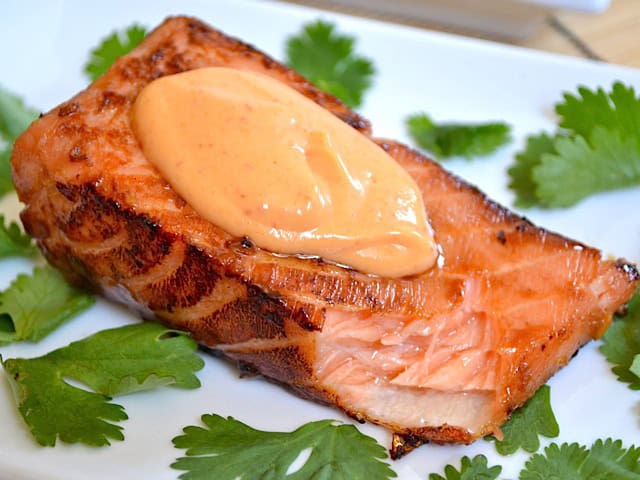 A piece of grilled salmon, inspired by classic salmon recipes, is topped with a dollop of creamy sauce and sits on a white plate, surrounded by fresh cilantro leaves.