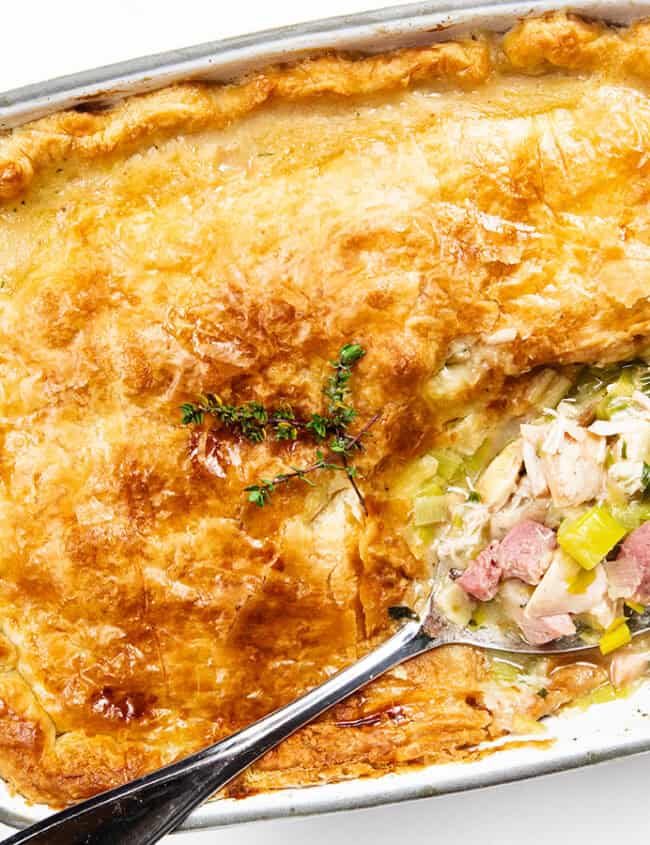 A baked dish with a golden-brown pastry crust partially removed, revealing a filling of chicken, ham, and leeks. Garnished with fresh thyme, it sits in a white rectangular casserole on a wooden board.