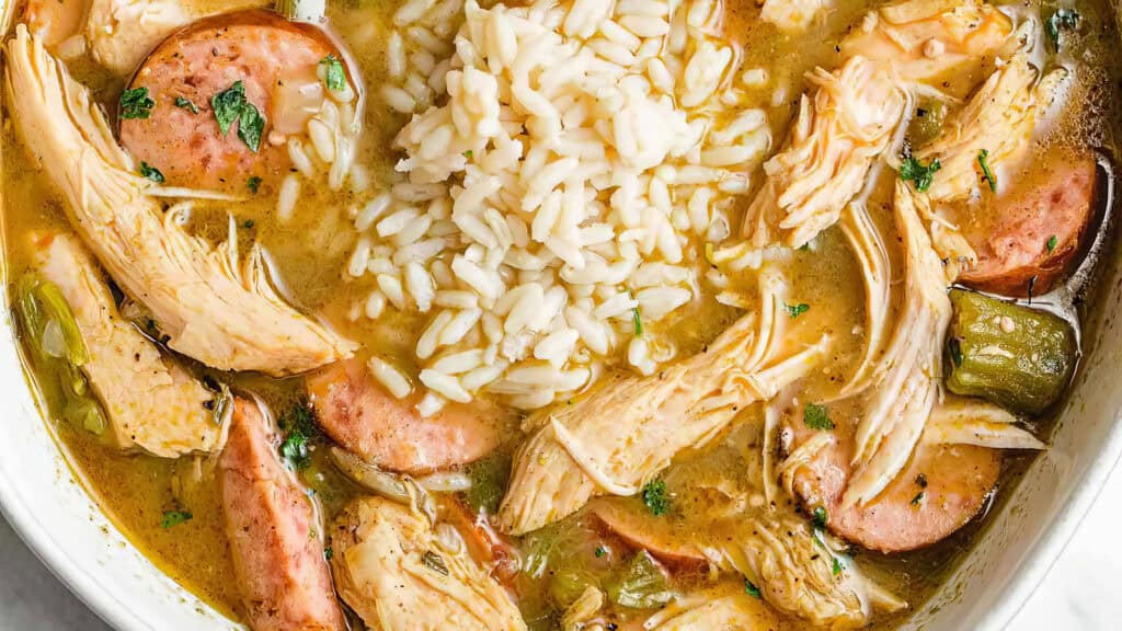 A bowl of gumbo featuring shredded chicken, sliced sausage, okra, and a scoop of white rice. The dish is garnished with herbs, set in a savory broth, capturing a hearty and comforting meal.