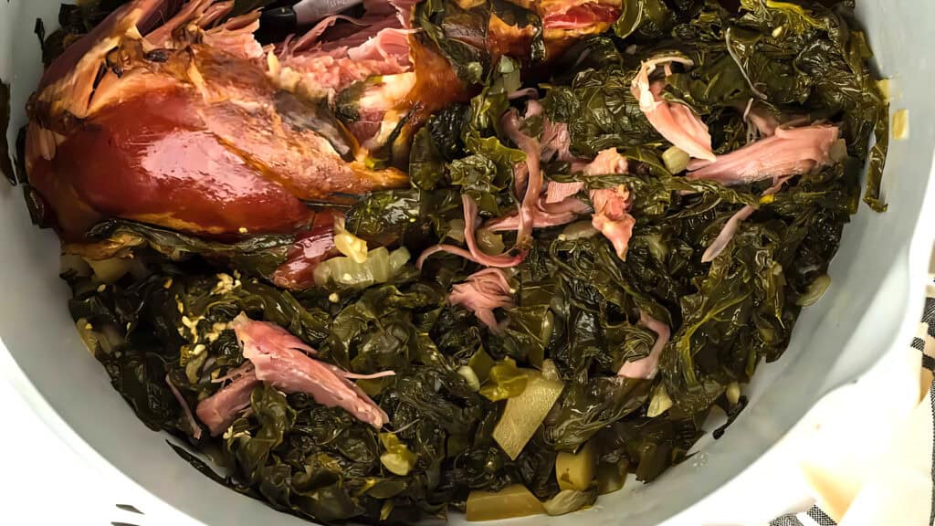 A close-up of a pot filled with cooked collard greens and pieces of smoked turkey. The greens are dark green and wilted, while the turkey is tender and pink, suggesting a savory, home-cooked dish.