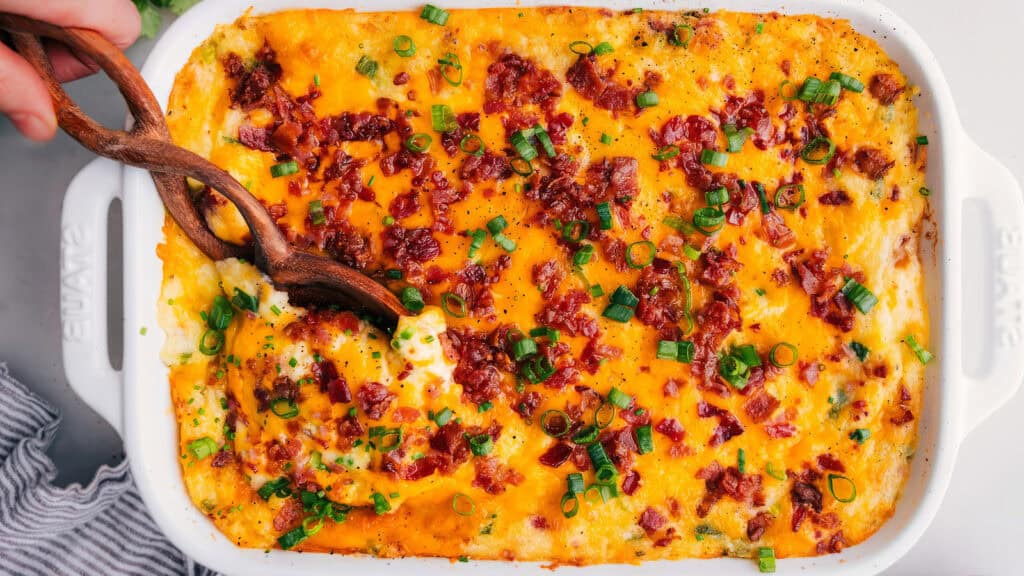 A rectangular baking dish filled with creamy mashed potatoes topped with melted cheddar cheese, crispy bacon bits, and chopped green onions. A wooden spoon is dipping into the dish, showing a serving scoop.