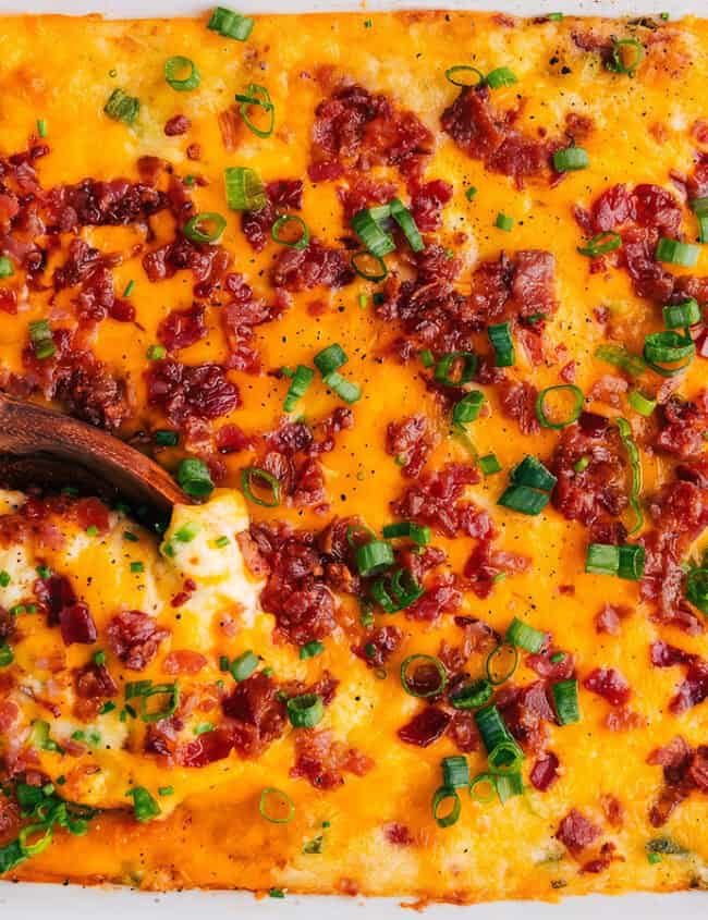 A rectangular baking dish filled with creamy mashed potatoes topped with melted cheddar cheese, crispy bacon bits, and chopped green onions. A wooden spoon is dipping into the dish, showing a serving scoop.