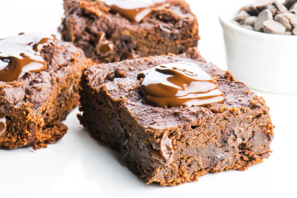 Close-up of three chocolate brownies from our favorite brownie recipes, topped with melted chocolate and arranged on a white surface. A small white bowl filled with chocolate chips is visible in the background. The brownies appear rich and chewy.