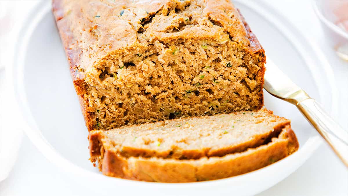 A loaf of freshly baked zucchini bread rests on a white plate. One slice is partially cut and lies at the front, revealing a moist and fluffy interior speckled with green zucchini pieces. A knife is placed beside the loaf.