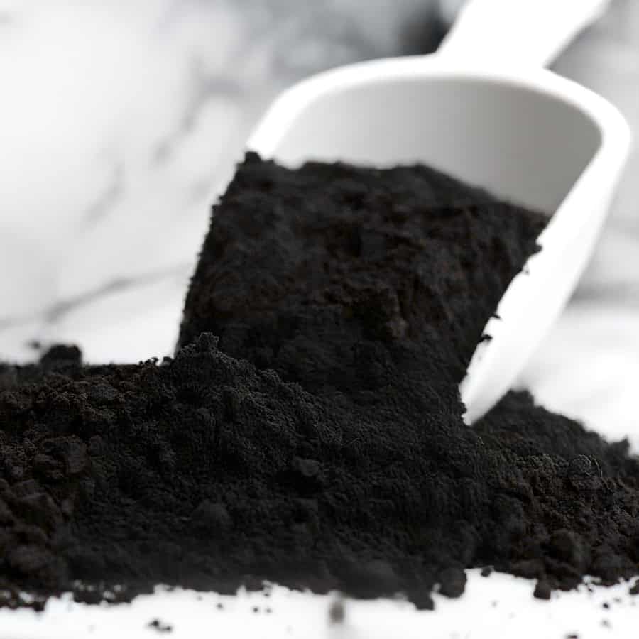 A white scoop holds a pile of fine black powder, reminiscent of rich Black Velvet Cake, spilling some onto a smooth surface. The background is softly blurred, highlighting the texture and evoking the luxurious feel of velvet.