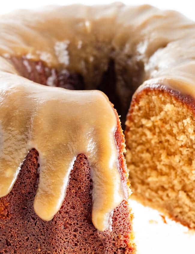 A bundt cake with a glossy caramel glaze drips down the sides. A slice is cut, revealing a moist, golden interior. The cake is displayed on a white background, highlighting its rich texture and appetizing appearance.