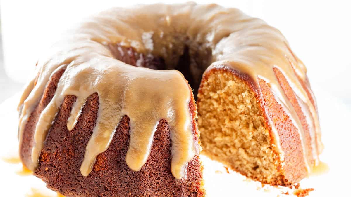 A bundt cake with a glossy caramel glaze drips down the sides. A slice is cut, revealing a moist, golden interior. The cake is displayed on a white background, highlighting its rich texture and appetizing appearance.