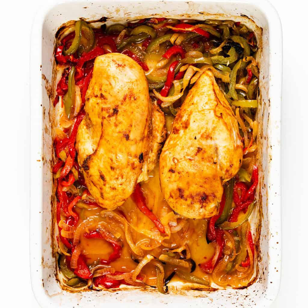 A baked dish featuring two seasoned chicken breasts surrounded by sliced red, green, and yellow bell peppers, topped with a sprinkle of cheese. The ingredients are in a white rectangular baking dish, cooked to a golden brown.