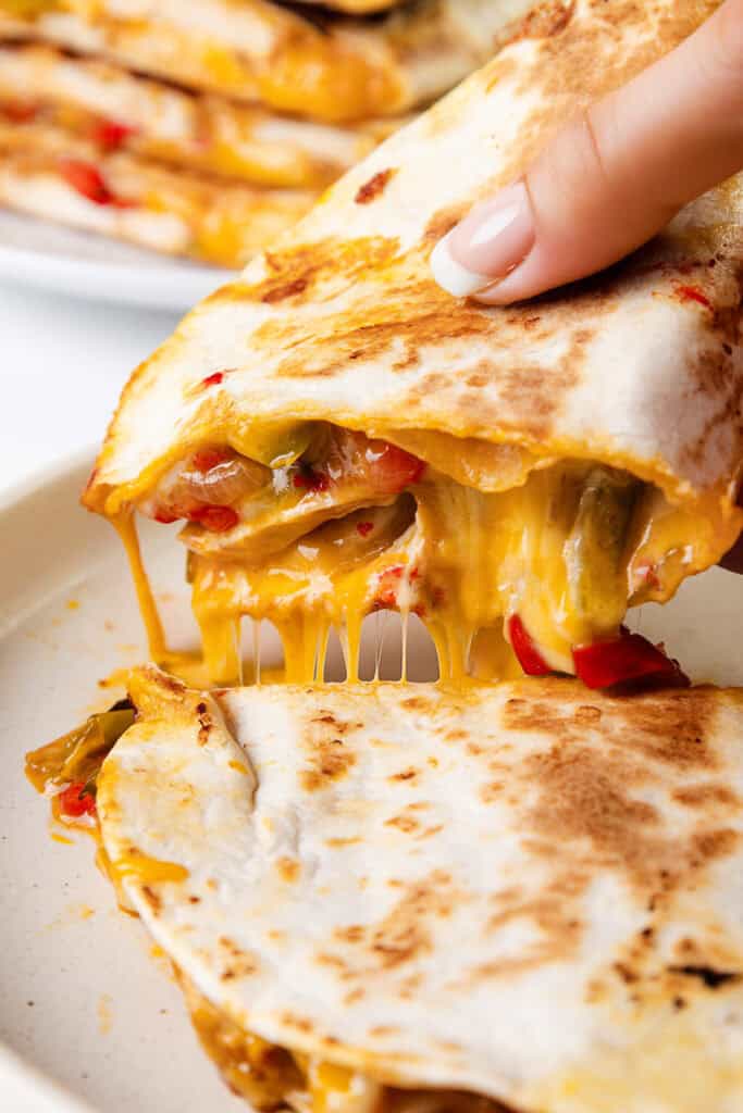 A hand pulling apart a cheesy chicken quesadilla reveals a luscious mix of melted cheese and perfectly cooked vegetables inside. The golden-brown quesadilla, showcasing vibrant bell peppers, is elegantly served on a white plate.