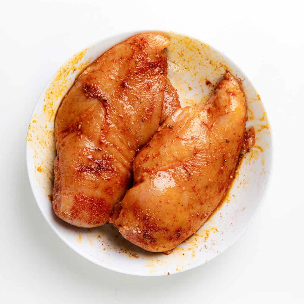 Two seasoned raw chicken breasts in a white bowl, coated with a reddish spice rub, are ready for cooking—a perfect start for savory quesadillas. Their vibrant color contrasts against the white background, promising an enticing dish ahead.